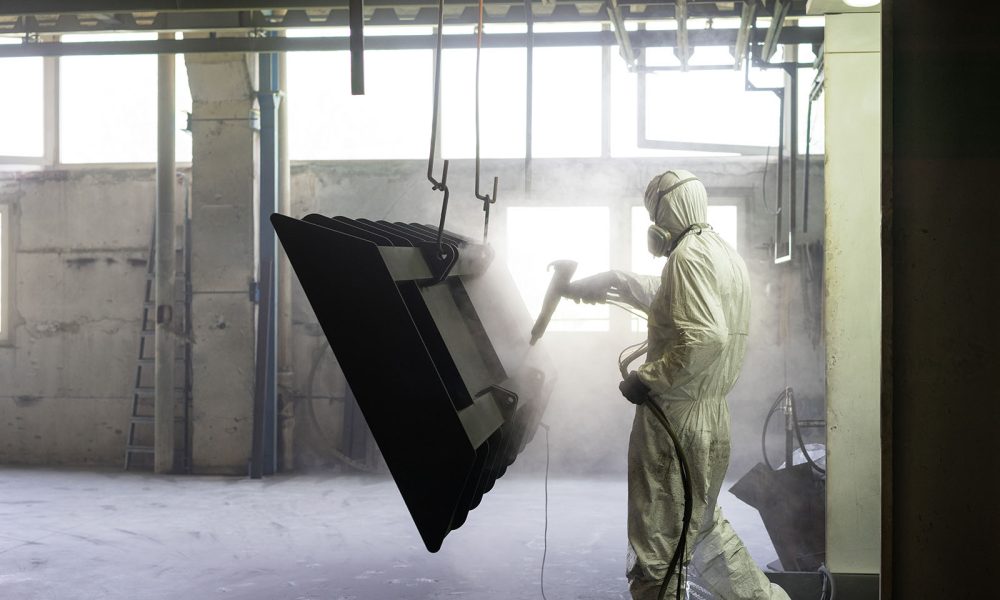 view of a worker wearing a full white protective suit and breathing mask, sand blasting a metal crate hung from a metal beam in the ceiling of an industrial hall